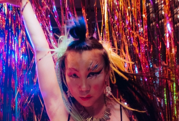 Queer to Stay thumbnail, image of asian person dancing in front of rainbow tinsel curtain