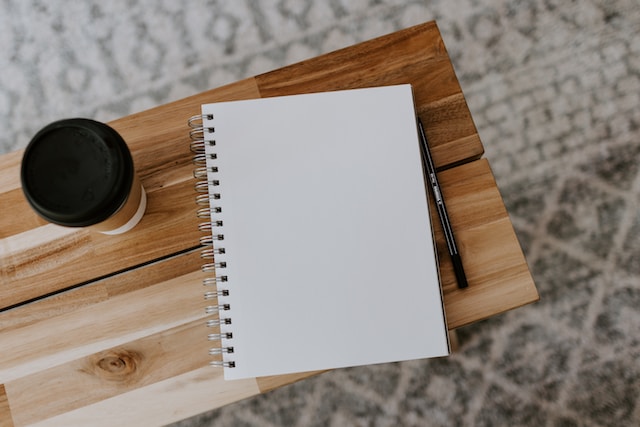 A cup of coffee, notebook, and pen sit together on a small wooden desk, seen from above, the grey and white pattern of the tile below is also visible.